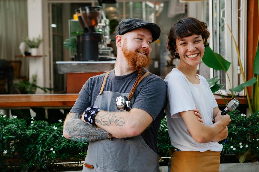 pexels-photo-6205478-6205478 Couple of smiling barista coworkers in casual outfit and aprons standing with portafilter near cafeteria in street near plants in daylight